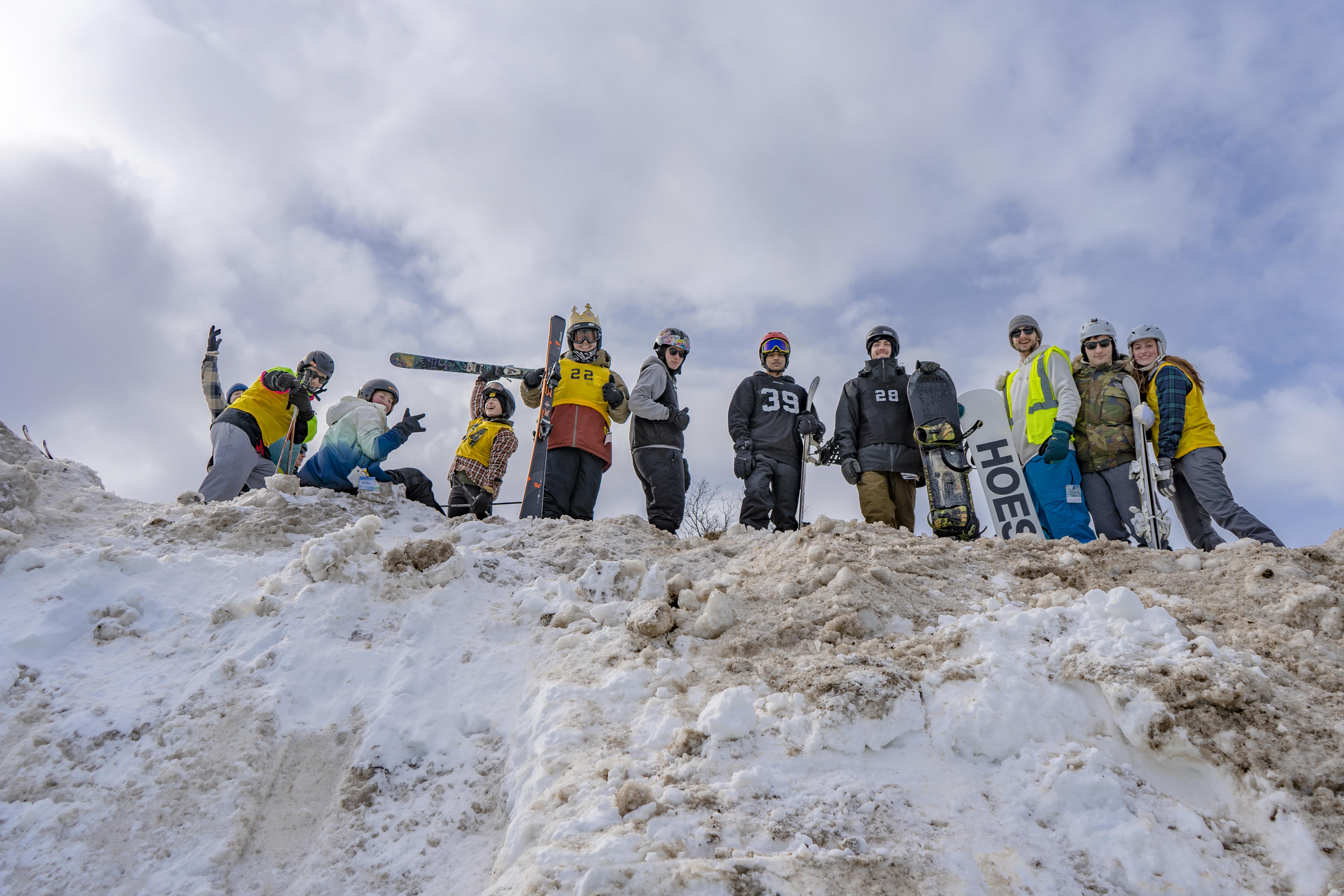 Skiers and snowboarders smile while standing atop a giant snow mound at a past Rail Jam