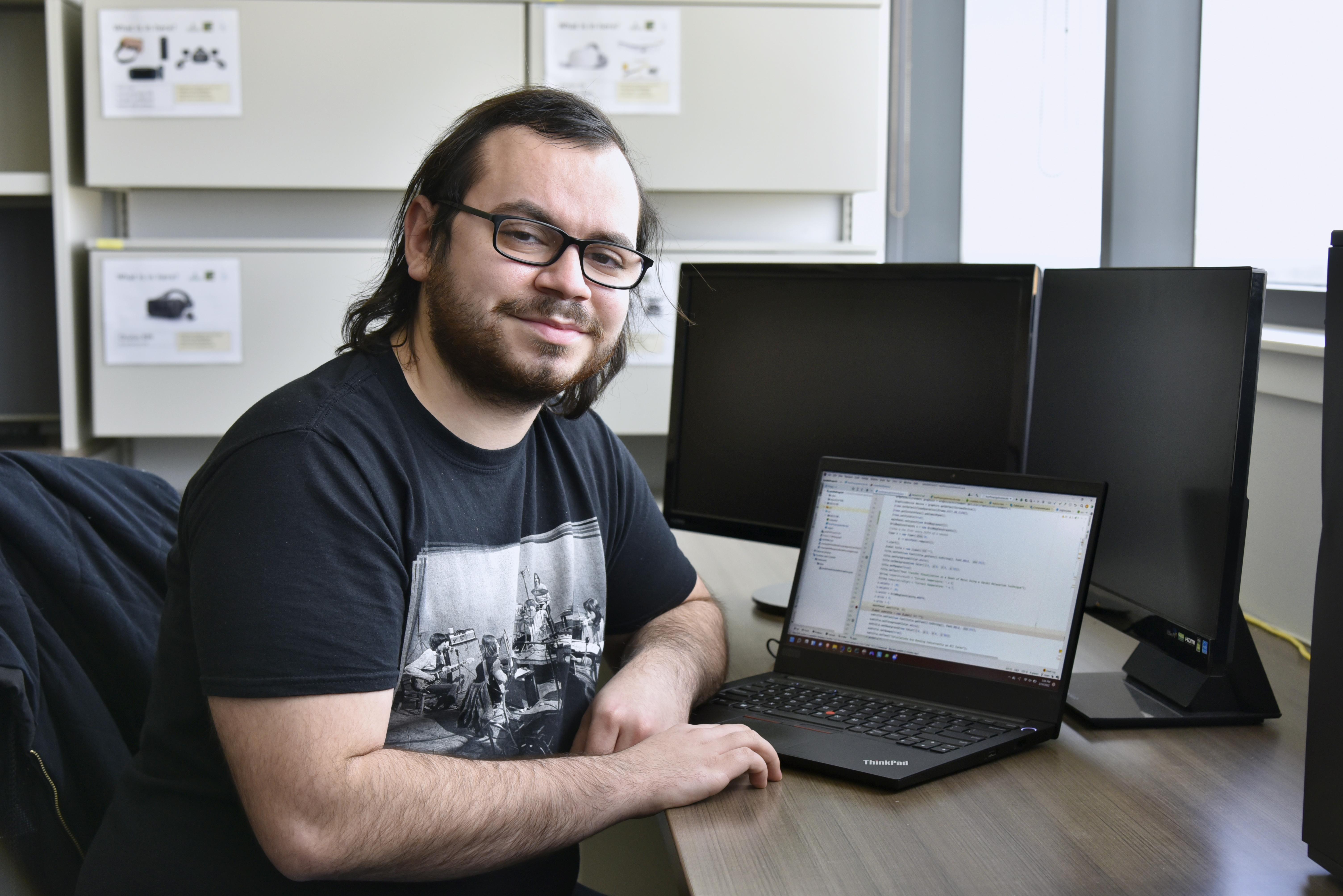 Computer science major Dominic Altamura sits in front of a computer, wearing a black t-shirt and black glasses. He has mid-length brown hair.