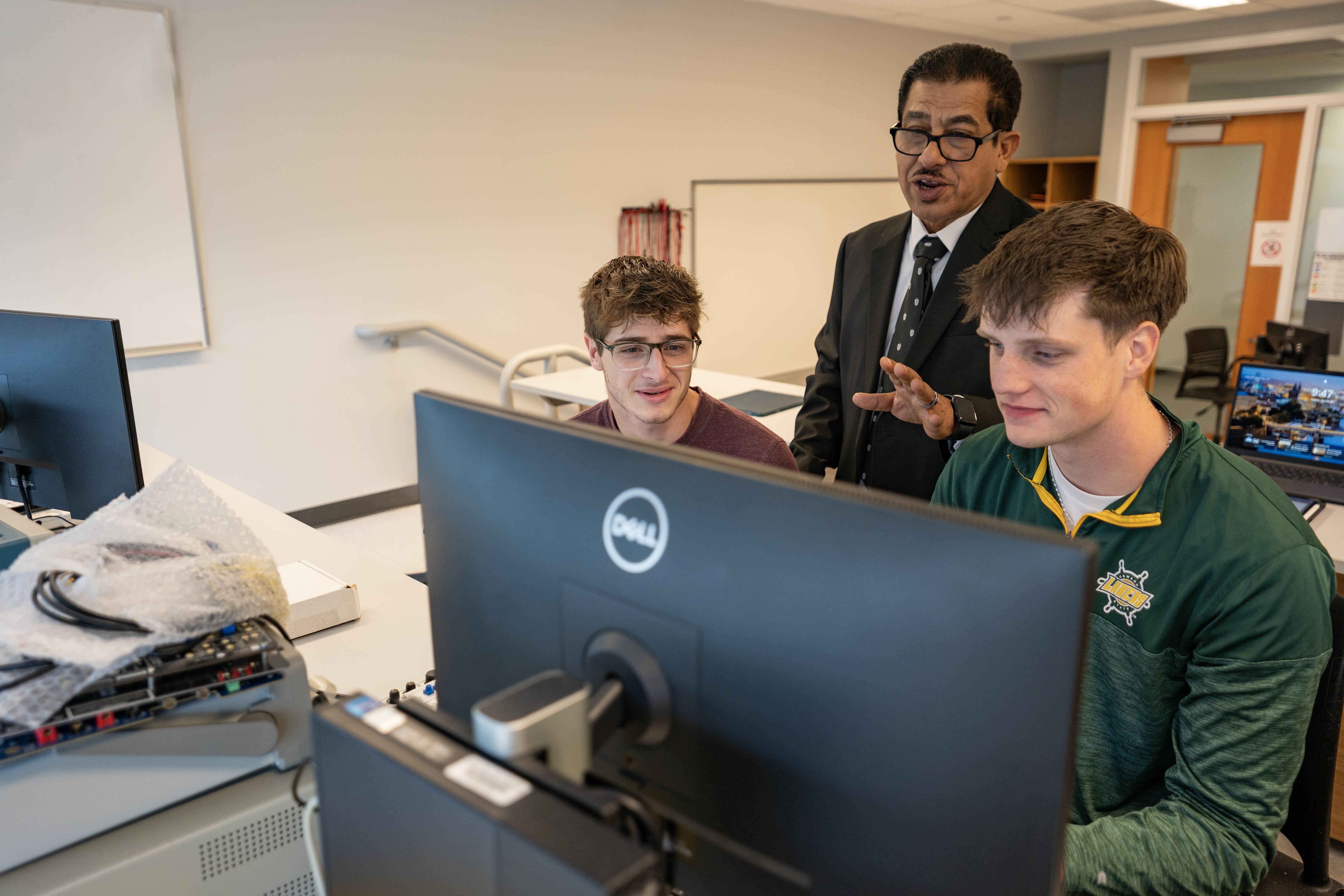A faculty member stands behind two SUNY Oswego engineering students as they work together at a computer in an electrical and computer engineering lab.