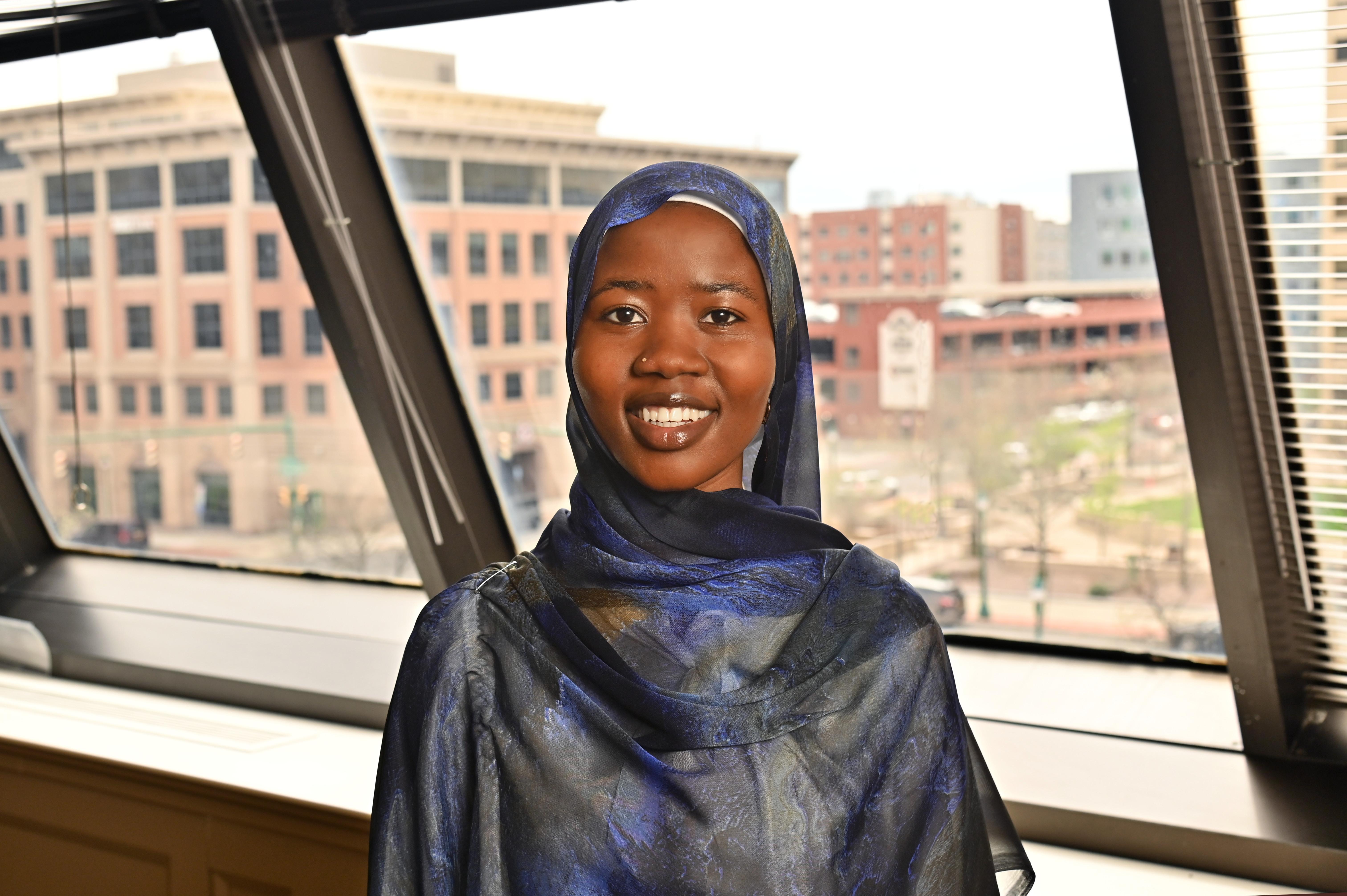 Student at SUNY Oswego at Syracuse smiles while posing indoors in front of large windows overlooking downtown Syracuse.