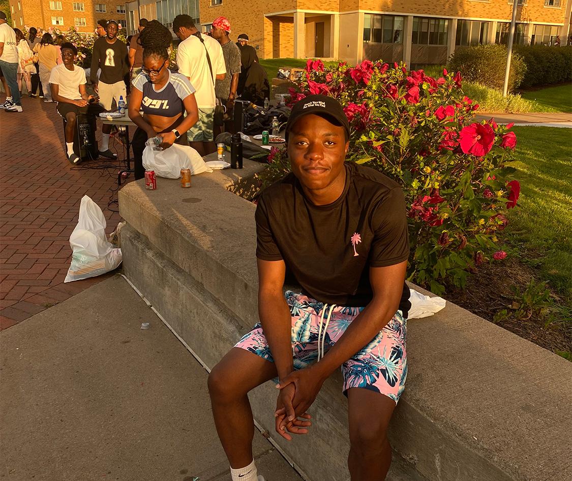 Antoine Okeke sits outside Hart Hall with the long line of students behind him waiting to try his menu for late night at Cooper Dining Hall. The sun is setting.
