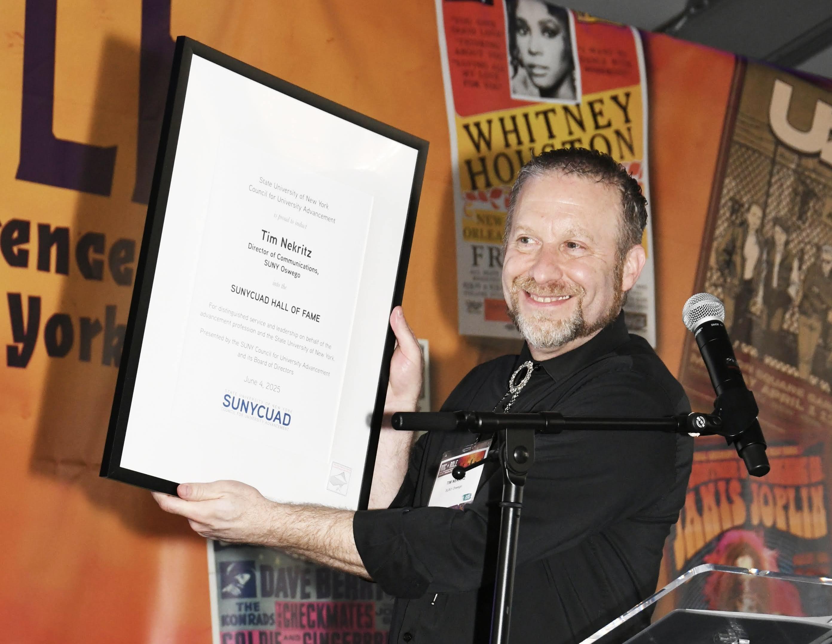 Tim Nekritz holds up his SUNYCUAD Hall of Fame Award on stage.