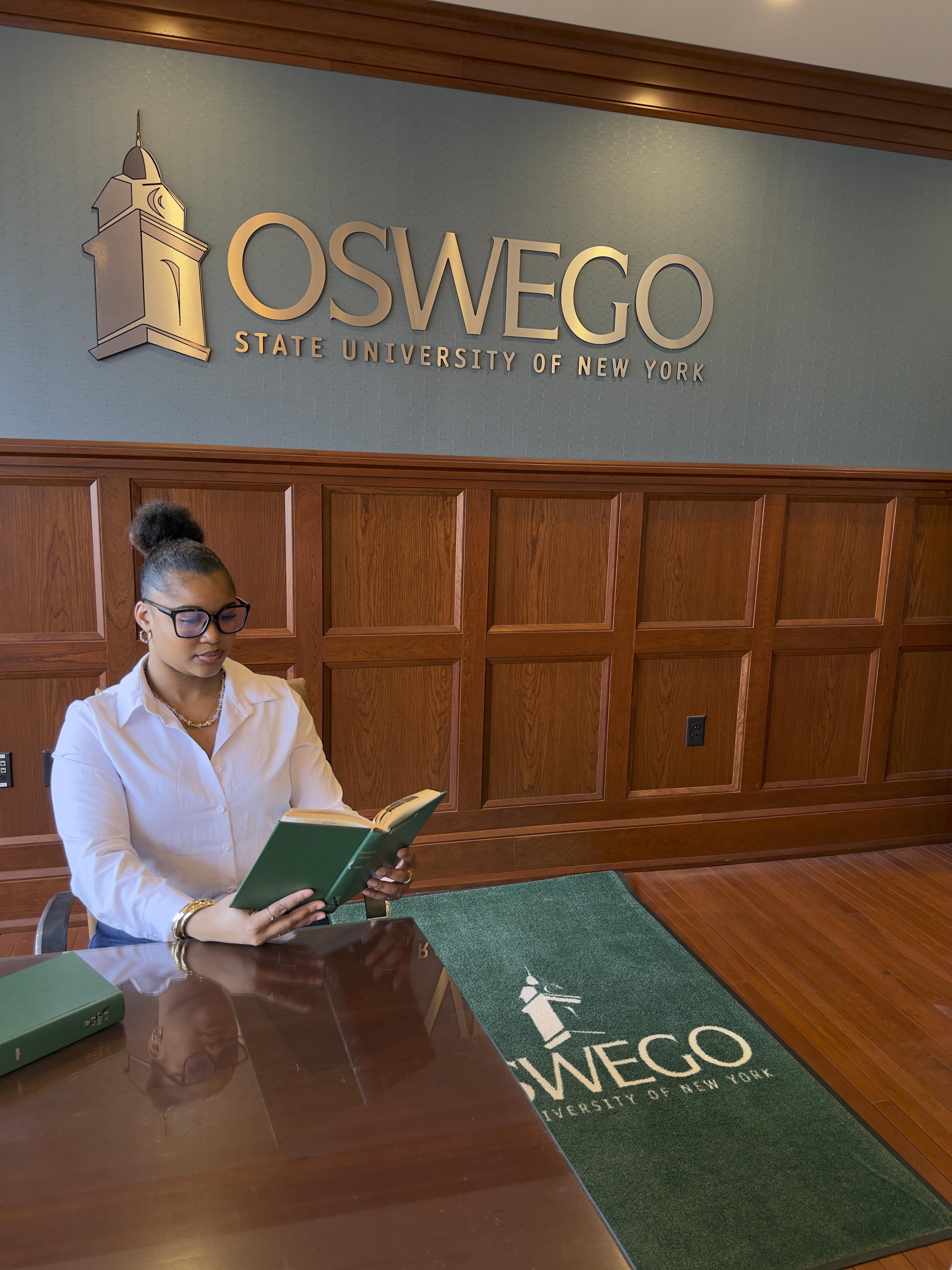 Maiya White reads a book while seated beneath SUNY Oswego signage at the university’s Syracuse campus.