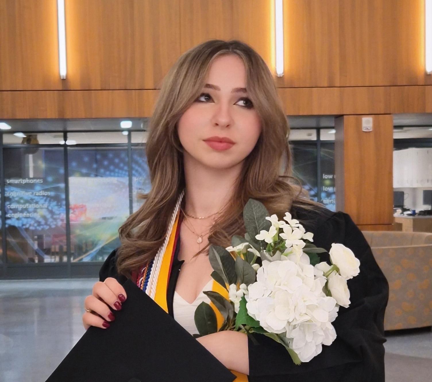 SUNY Oswego graduate Samantha Brown wearing a black cap and gown with honor cords and medals, holding a bouquet of white flowers while standing inside a modern campus building.