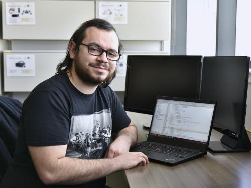 Computer science major Dominic Altamura sits in front of a computer, wearing a black t-shirt and black glasses. He has mid-length brown hair.