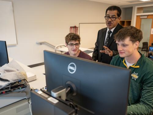 A faculty member stands behind two SUNY Oswego engineering students as they work together at a computer in an electrical and computer engineering lab.