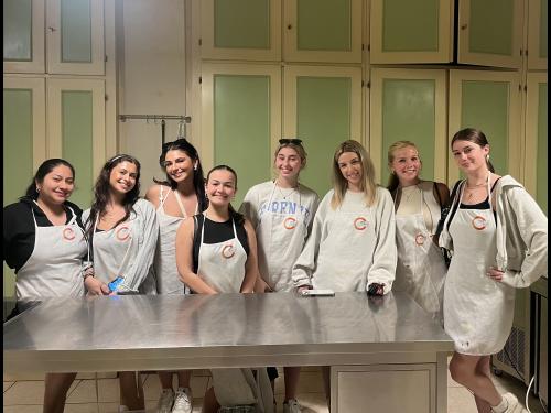 Students in the "Travel and Tourism" study abroad course pose for a picture at their cooking class held at Chefactory Cooking Academy in Florence, Italy. The group is all wearing white aprons with the company logo.
