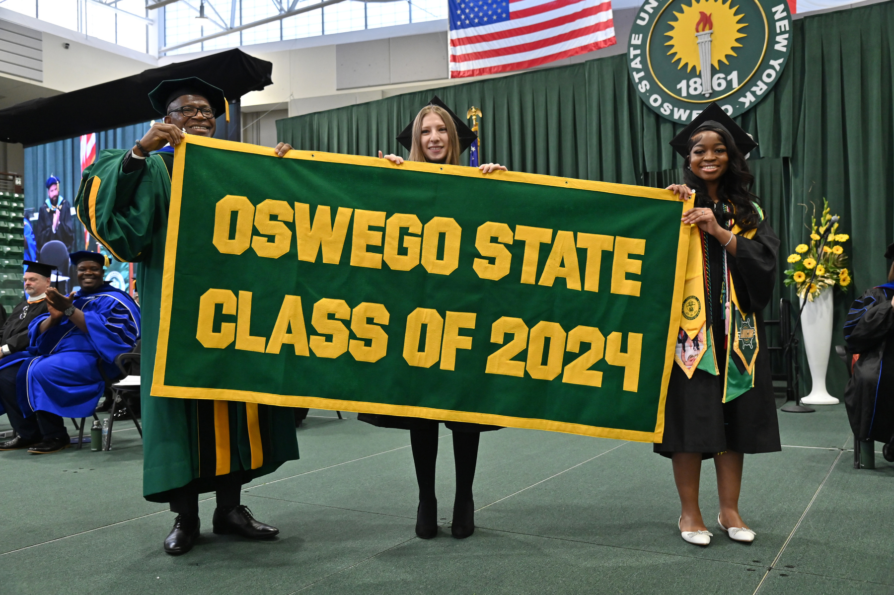 In the traditional alumni banner presentation ceremony at Commencement, from left, President Peter O. Nwosu receives the banner from graduate presenter Hayley Weiner (center), earning a master’s in higher education leadership, and Josephine Taylor, earning a bachelor’s in journalism.