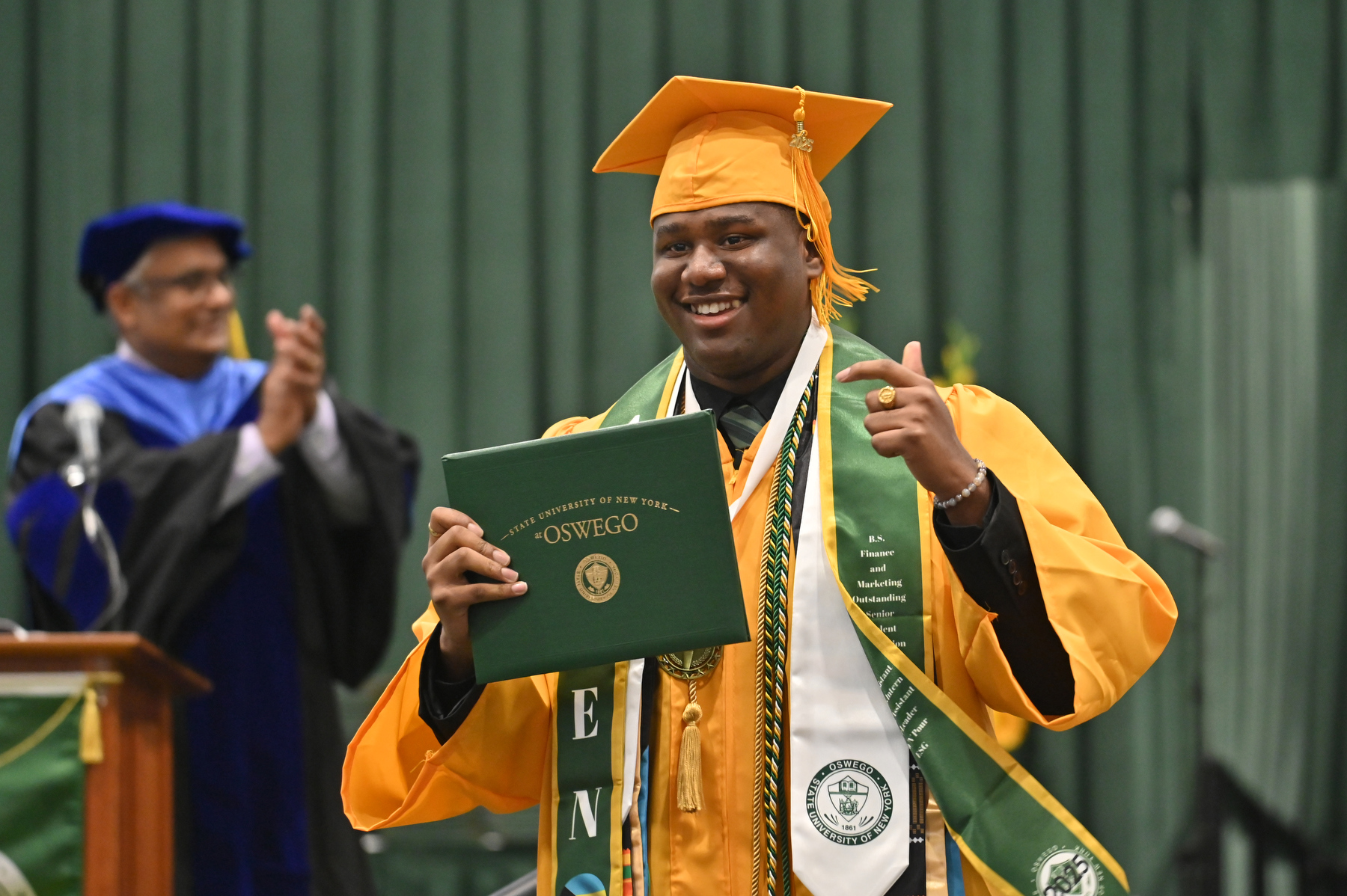 Aiden Wilson – Student Association president 2024-25 and the Oswego Alumni Association’s Outstanding Senior – walks across the platform to cheers from the audience, receiving degrees in finance and marketing. Pictured in background in Prabakar Kothandaraman, dean for the School of Business.